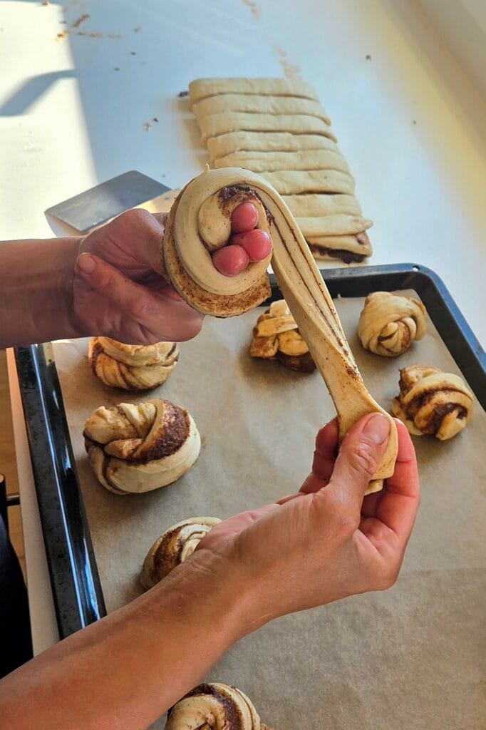 Hands twisting a strip of cardamom dough into a cinnamon knot over a baking sheet