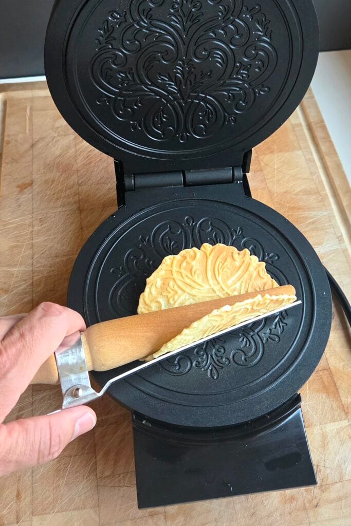 Rolling a hot krumkake around a wooden cone roller beside the open iron