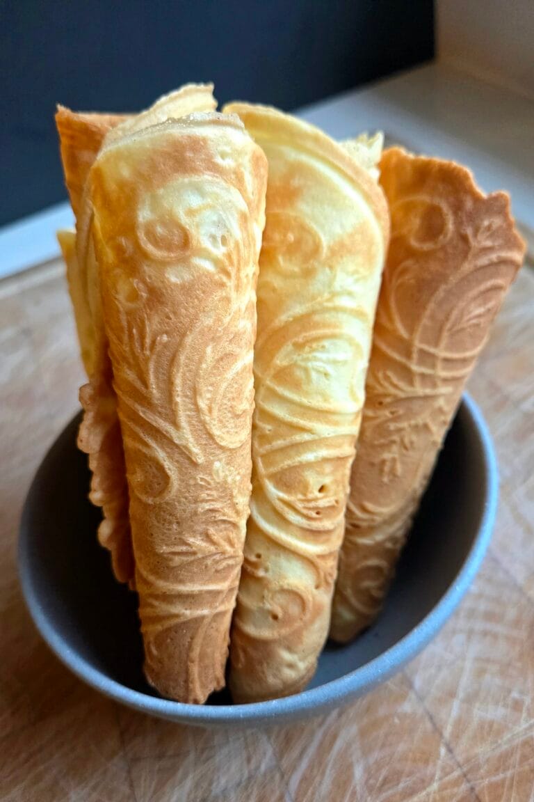 Krumkake cones stacked upright in a bowl, showing the rosemaling pattern pressed into each Norwegian waffle cookie