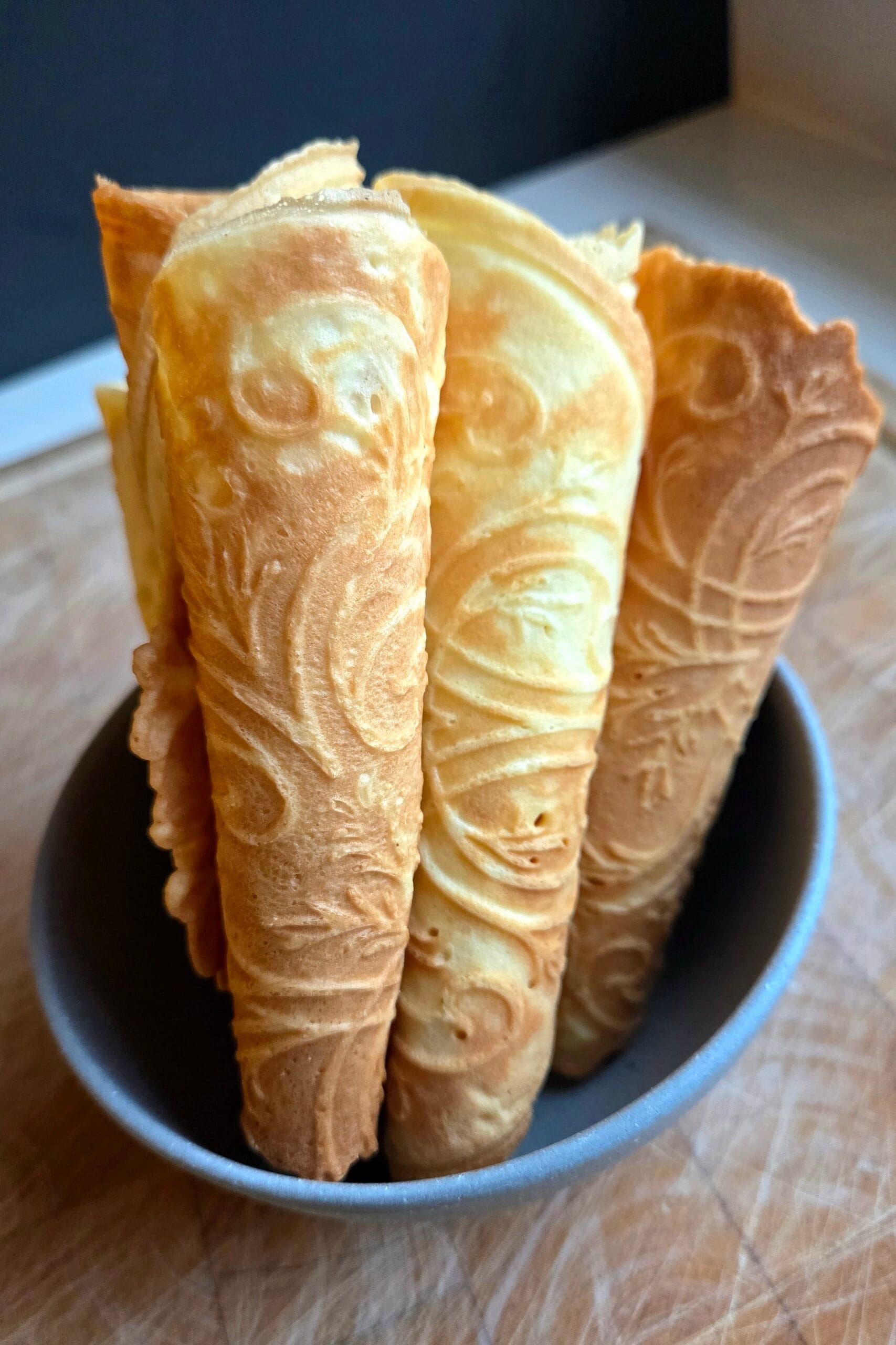 Krumkake cones stacked upright in a bowl, showing the rosemaling pattern pressed into each Norwegian waffle cookie