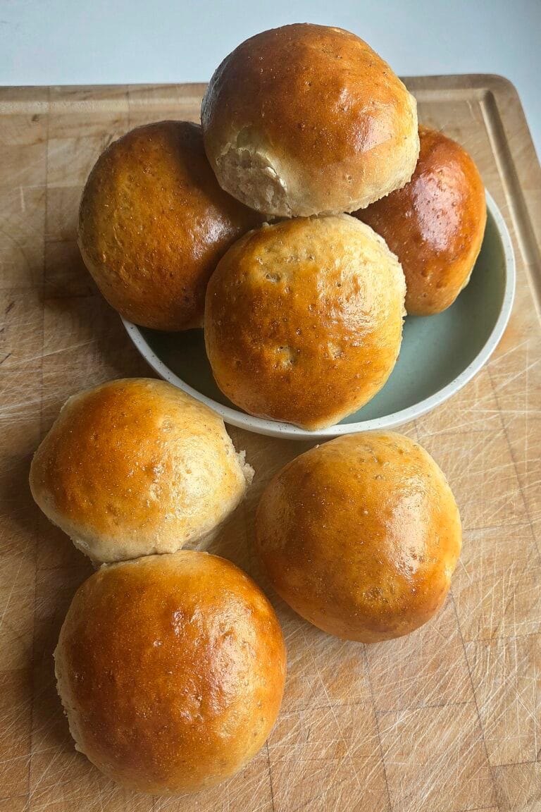 Golden Norwegian rye buns stacked on a wooden board with a green bowl