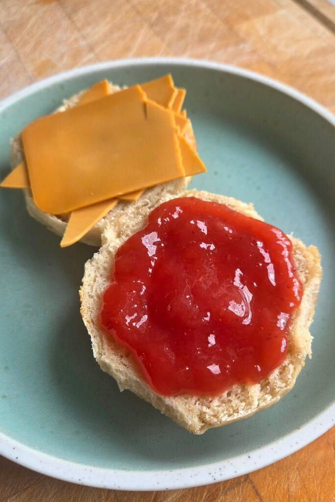 Split Norwegian rye buns on a green plate with brown cheese and strawberry jam