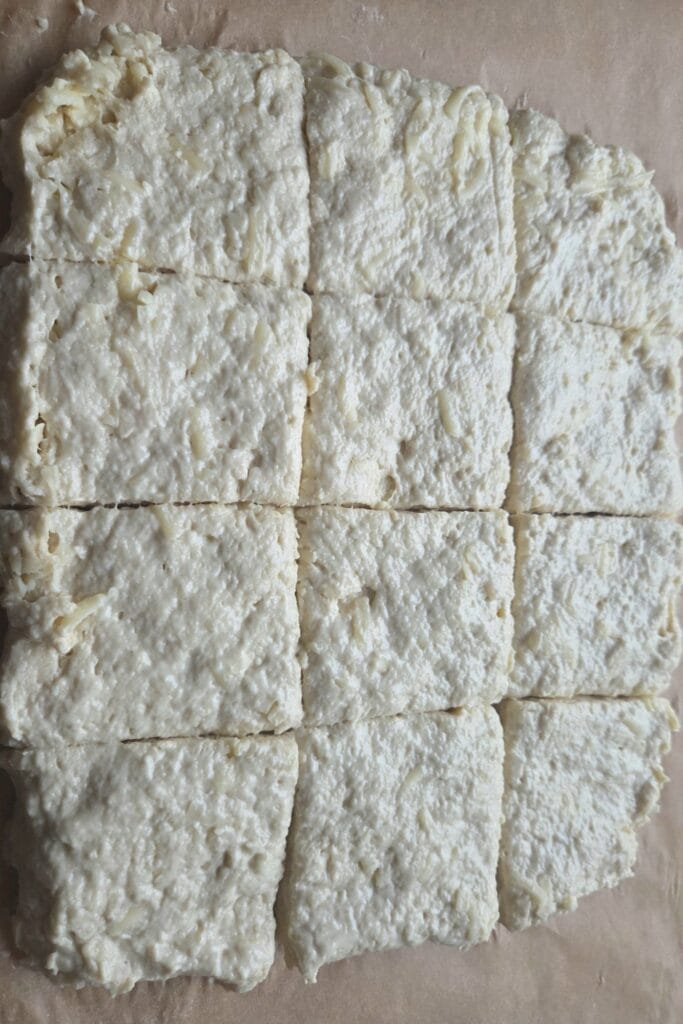 Cheese scone dough patted into a square and cut into 12 pieces on parchment paper