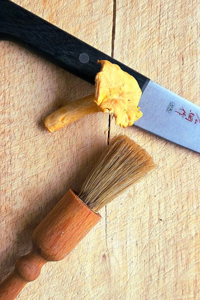 A paring knife and a mushroom brush on a wooden board with a trimmed chanterelle