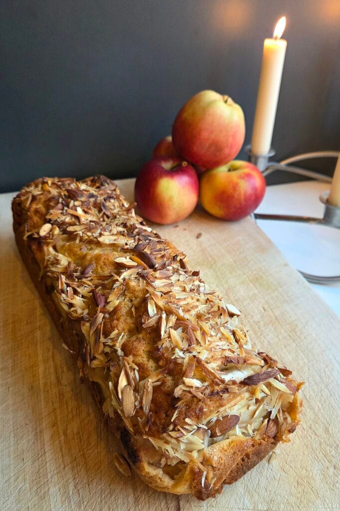 Whole Norwegian apple cake on a wooden board topped with toasted sliced almonds, with red apples and candlelight in the background