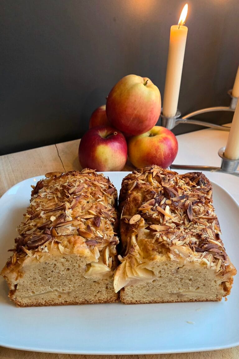Norwegian apple cake sliced open on a white plate, showing the apple layer inside and a toasted almond topping, with fresh apples and a lit candle behind