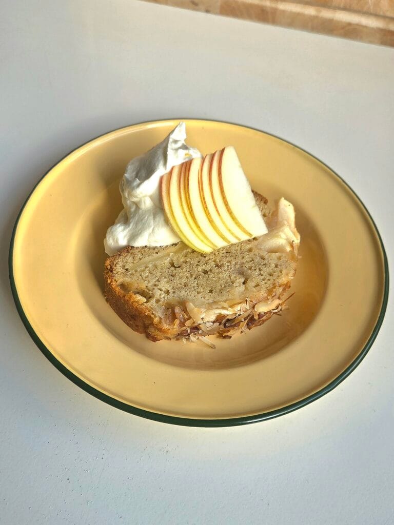 A slice of Norwegian apple cake on a yellow plate with whipped cream and thin slices of fresh apple fanned on top