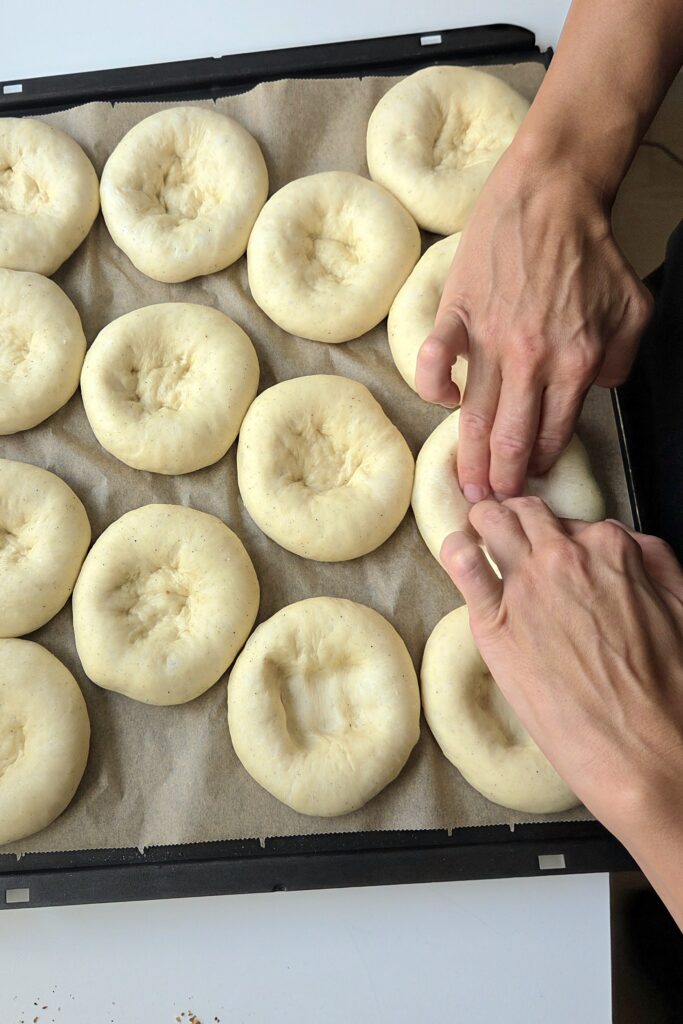 Fingertips pressing a wide, shallow well into a skolebrød bun, leaving a raised rim