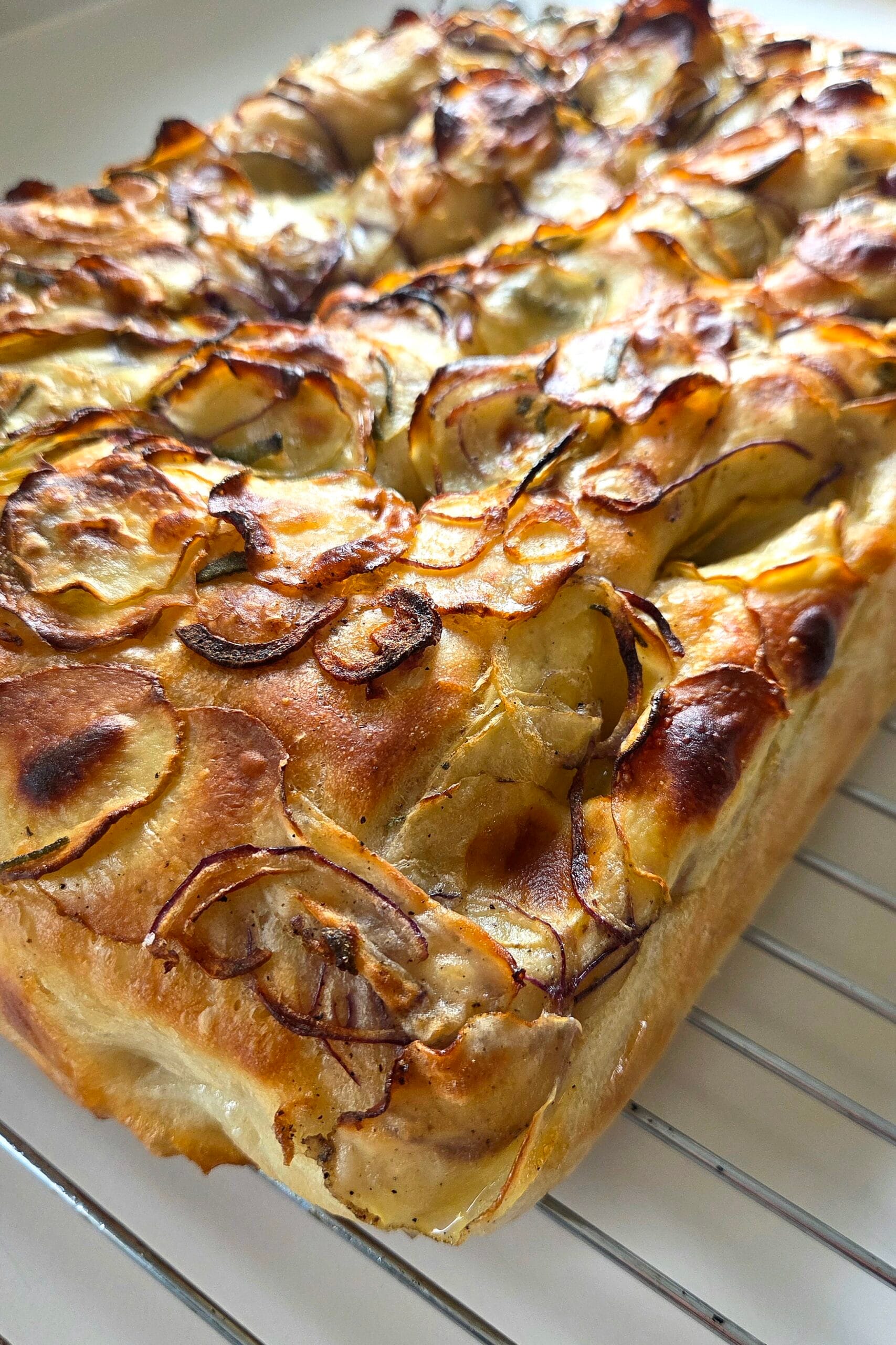 Potato focaccia with red onion and sage cooling on a wire rack, topped with crisp golden potato slices