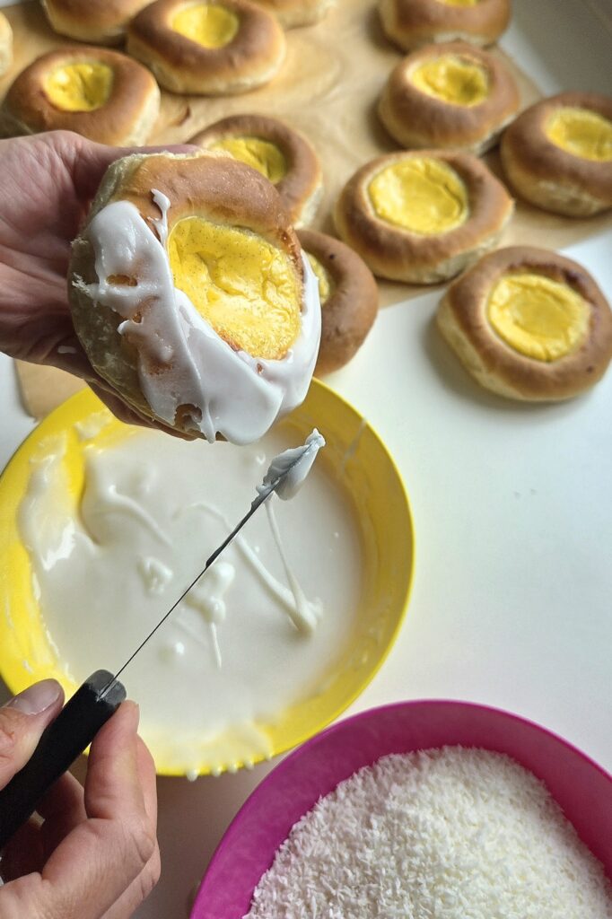 Spreading white glaze around the custard of a baked skolebrød with a small spatula, with bowls of glaze and desiccated coconut ready for dipping