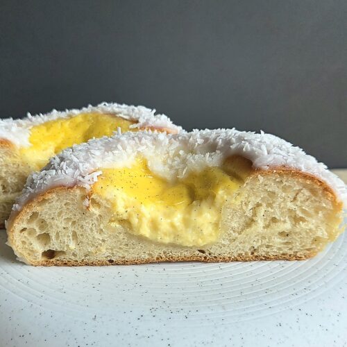 Skolebrød cut in half on a white plate, showing the soft cardamom crumb and vanilla custard filling under a coconut-covered glaze