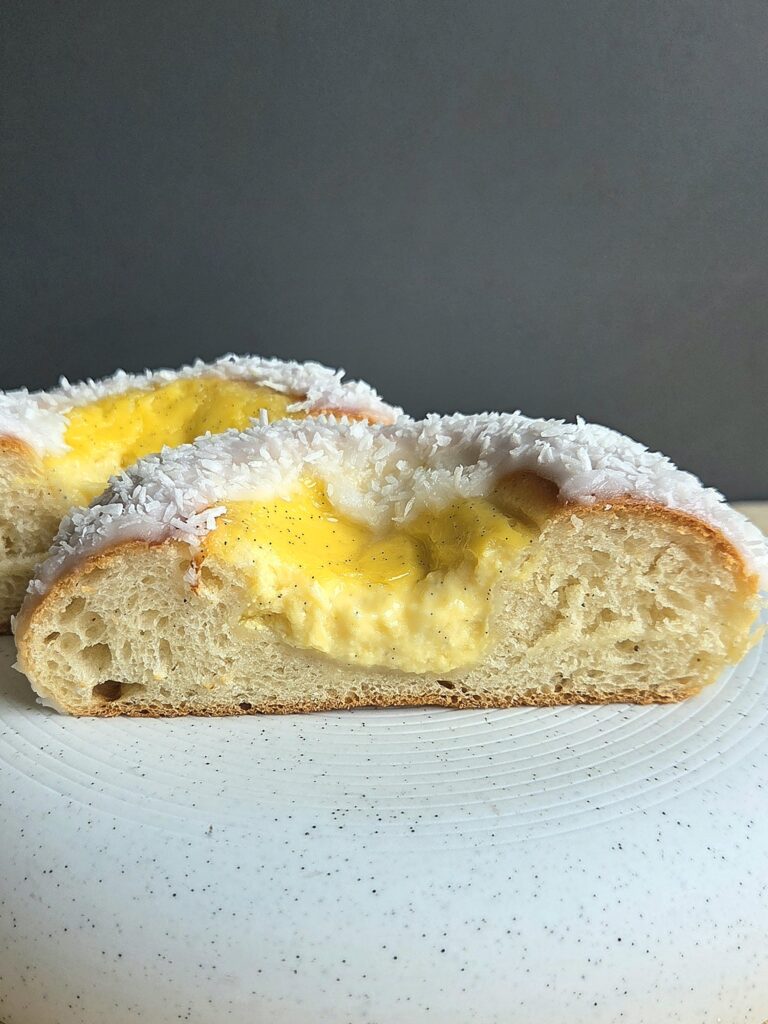 Skolebrød cut in half on a white plate, showing the soft cardamom crumb and vanilla custard filling under a coconut-covered glaze
