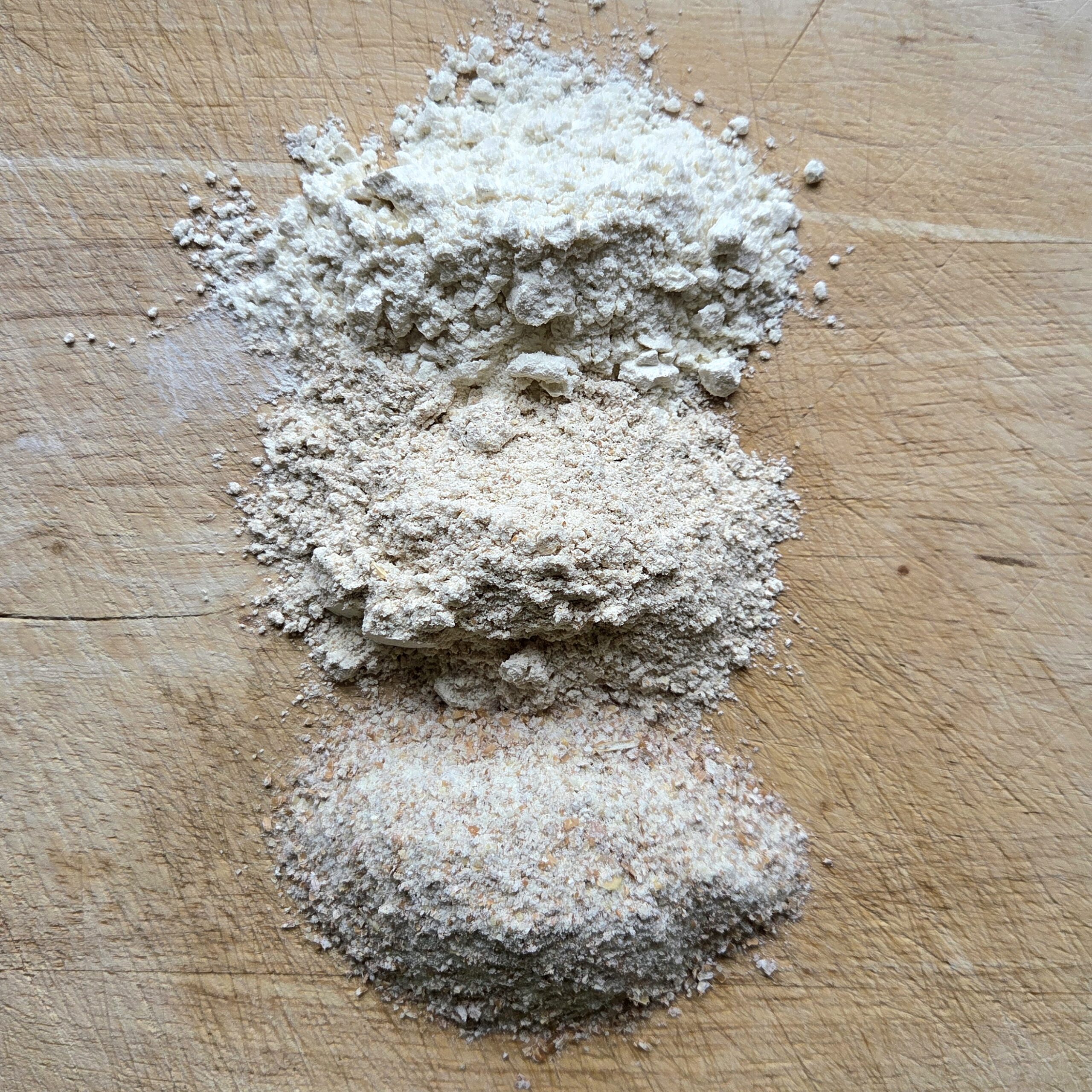 Three types of wheat flour compared on a wooden board — sifted, stone-ground and whole grain