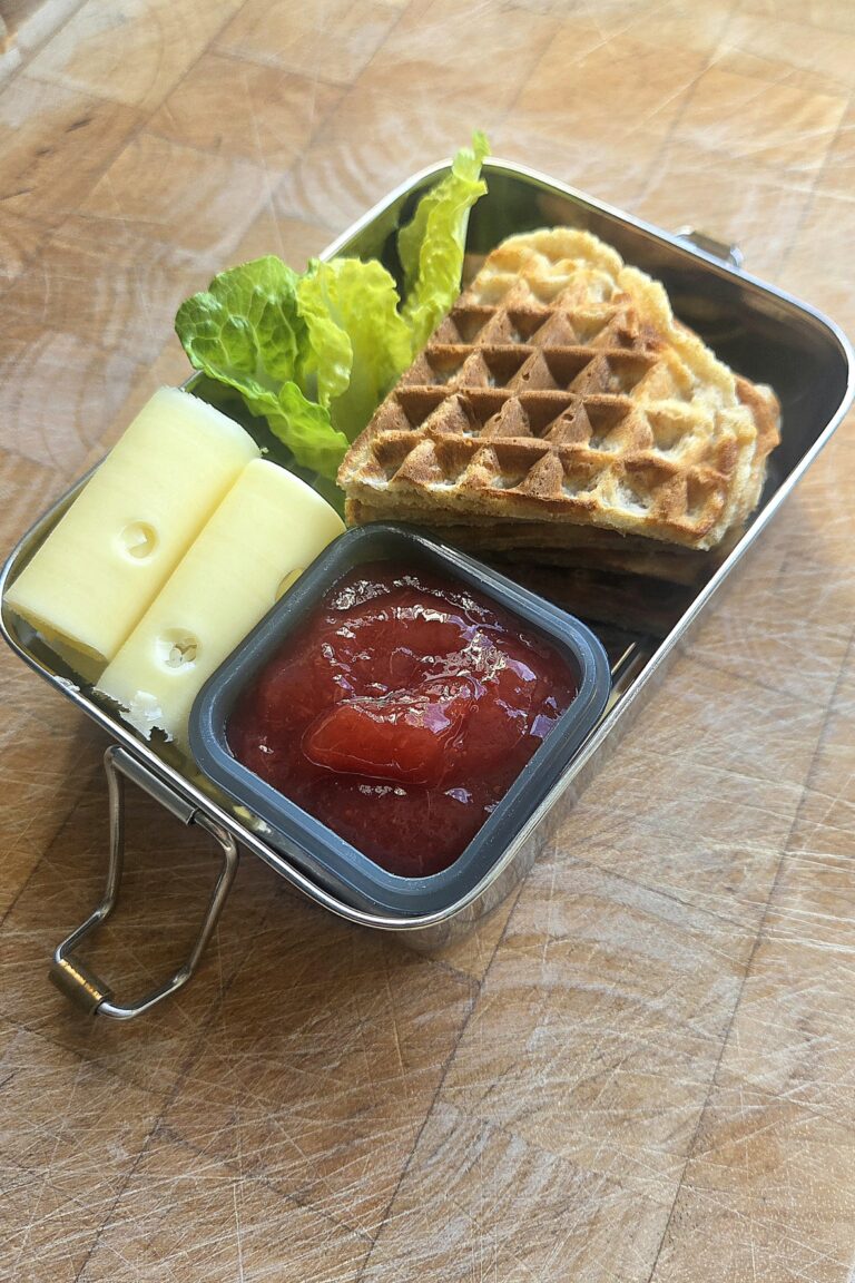 Steel lunchbox packed with whole wheat waffle hearts, rolled cheese slices, romaine and a small cup of red jam