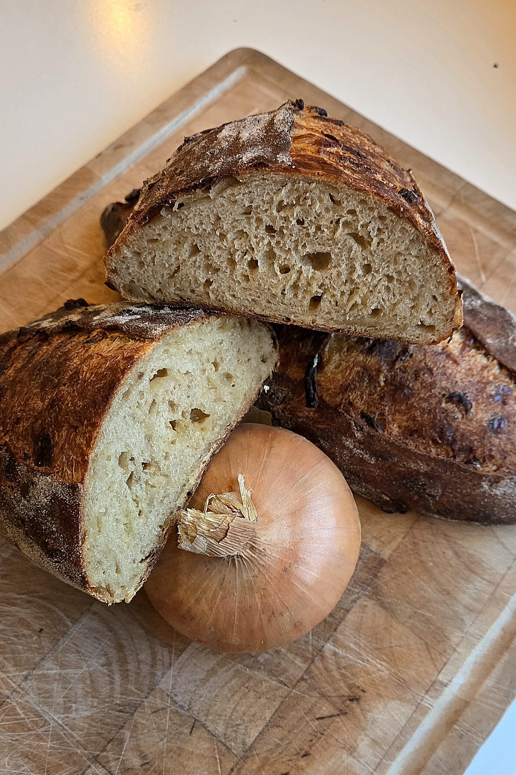 French onion soup sourdough cut into halves and stacked on a wooden board beside a whole yellow onion, with dark onion pieces in the crust